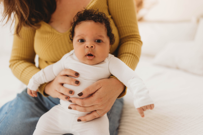 Mother gently holding baby in white clothing while sitting on bed